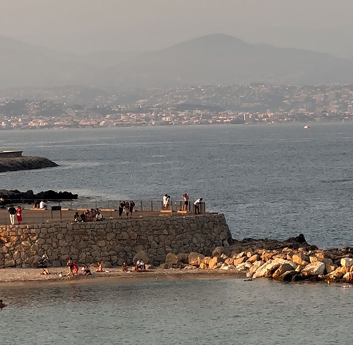 People relax on a rocky ledge overlooking a calm sea at sunset, with distant mountains and a cityscape in the background.