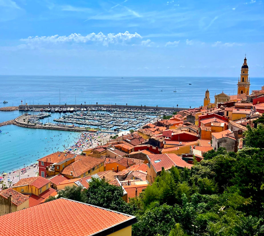 Menton coastal town with red-roofed buildings, bustling beach, marina with boats, and a blue sea under a clear sky. Vibrant and lively scene.