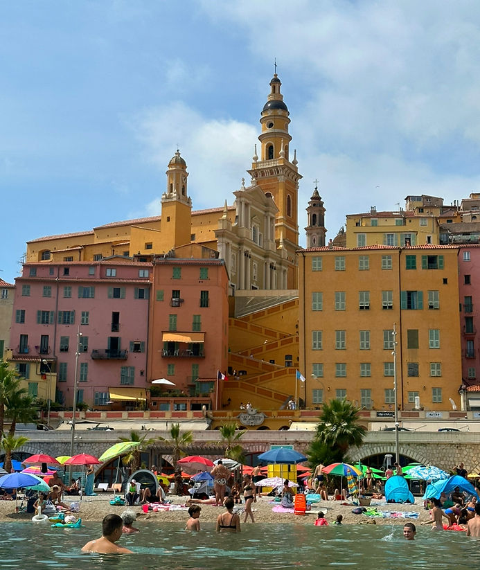 Crowded beach with colorful umbrellas and people in the water. Background shows pastel buildings and a large church. Bright and lively scene.