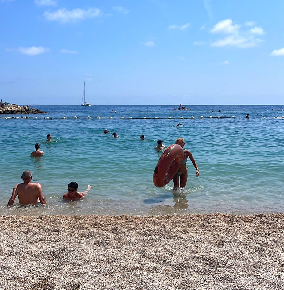 People enjoy a sunny beach; some swim while a woman enters the sea with a float. Blue sky, sailboat, and distant buoys are visible.