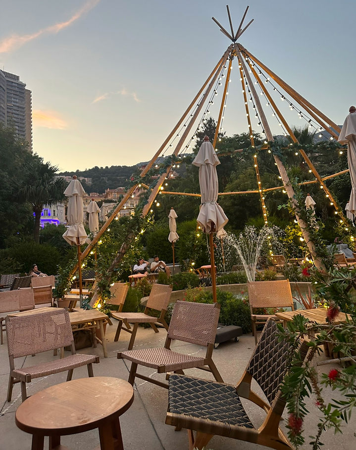 Outdoor seating area at sunset with empty chairs and tables. A teepee structure adorned with lights, fountain, and greenery decorate the space.