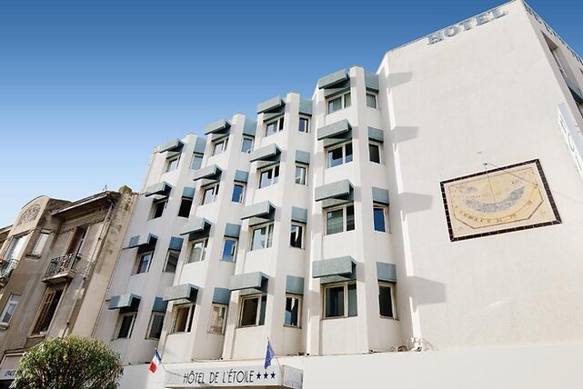 White hotel building with blue accents under clear sky; sundial on wall. Sign reads "Hôtel de l'Étoile." European flags displayed.