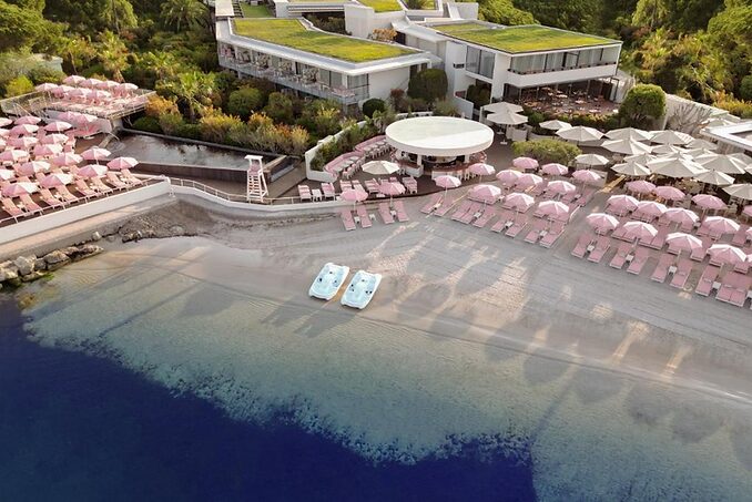 Aerial view of a beach resort with pink umbrellas and lounge chairs. Two pedal boats on the sandy shore, lush greenery surrounds modern buildings.