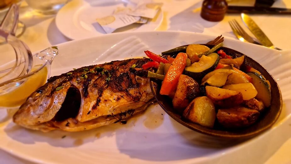 Grilled fish and roasted vegetables on a white plate with a sauce jug. Warm, inviting setting with soft lighting and neutral tones.