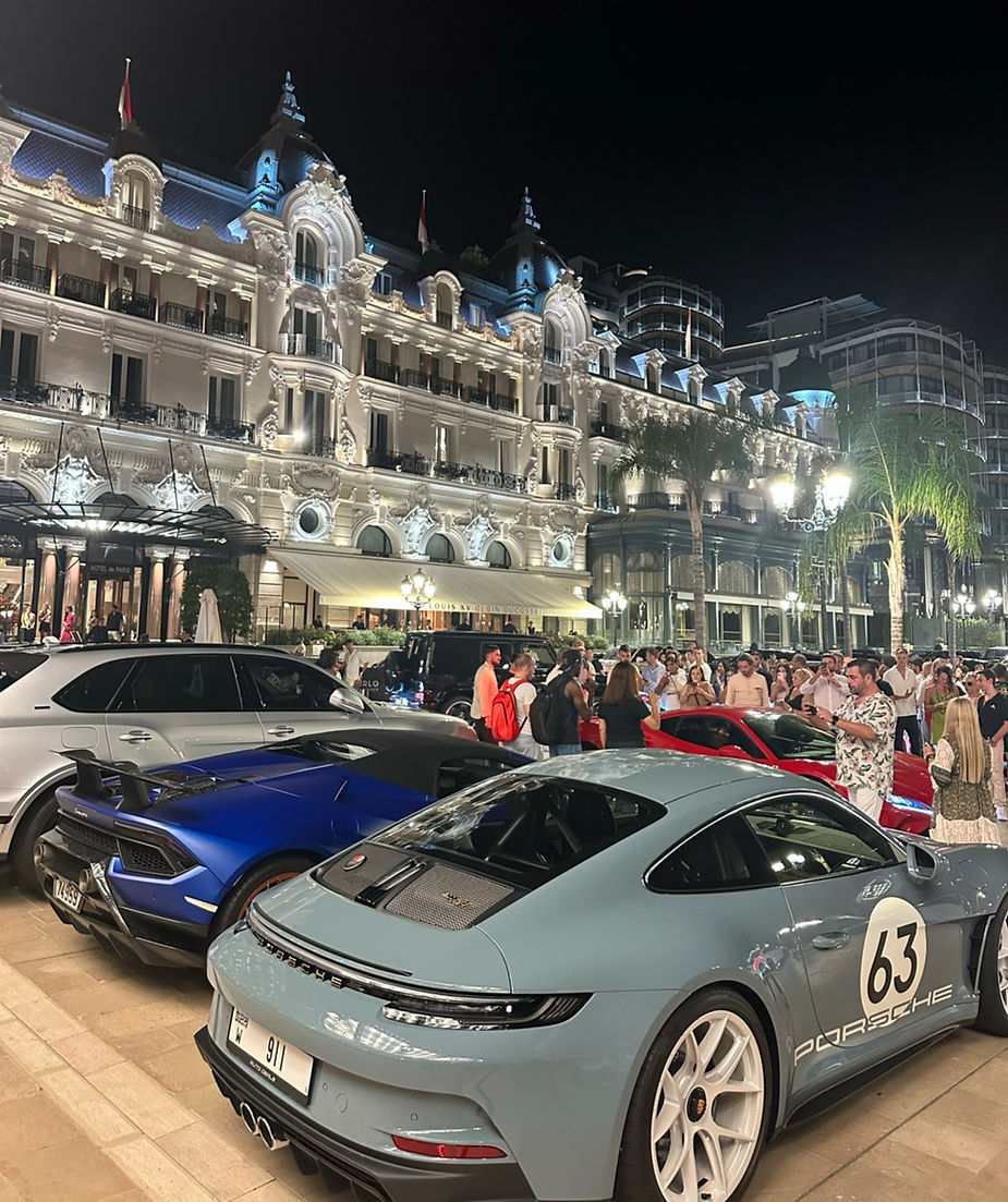 Luxury cars parked outside Casino Monte Carlo. A crowd gathers around, creating a lively, upscale atmosphere.