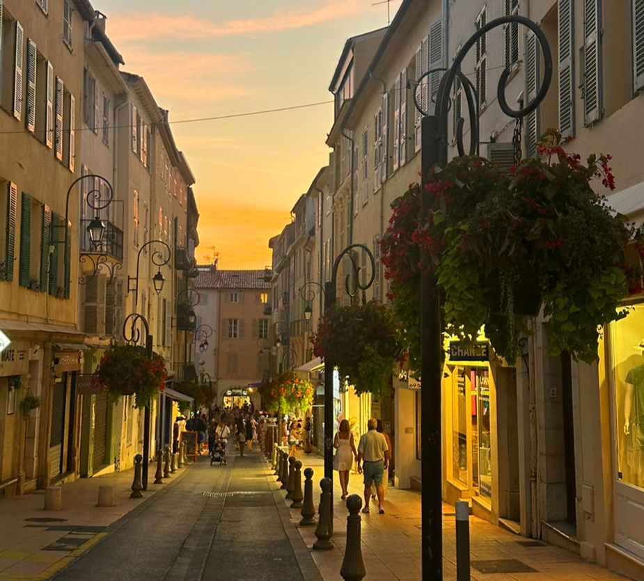 Street at sunset with people walking, flanked by lit shops and hanging plants. Warm glow in the sky, creating a cozy, inviting atmosphere.