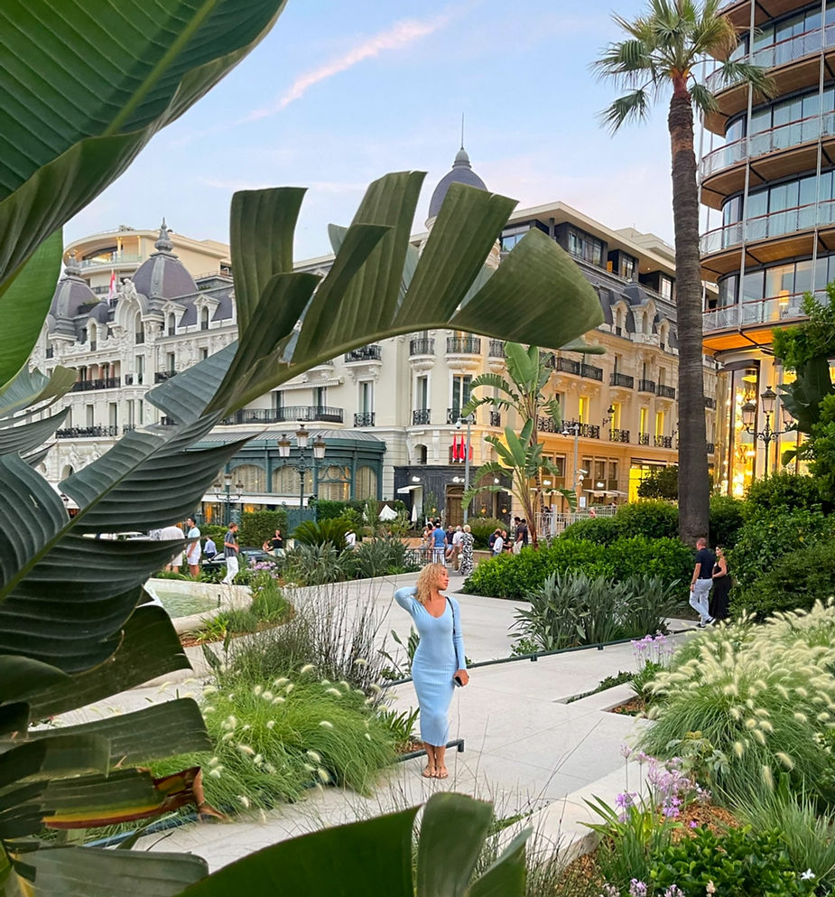 Blonde curly haired girl in blue tight dress stands in a lush garden, surrounded by tropical plants. Elegant buildings are in the background, creating a serene evening scene.