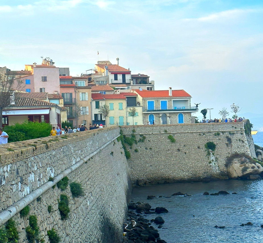 Coastal town with colorful buildings and a stone wall; people walk along the wall. Sea and rocky shore in the foreground, blue sky above.