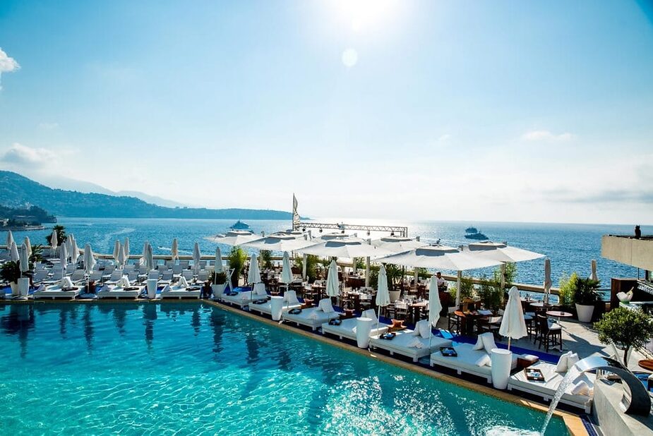 Luxurious poolside resort with white loungers and parasols overlooking a calm blue sea, mountains in the background. Sunny and tranquil atmosphere.