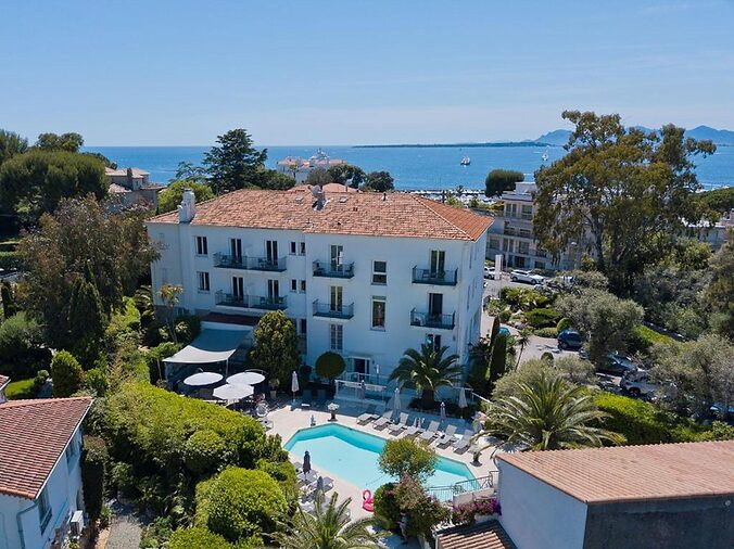 Aerial view of a white hotel with a red roof, surrounded by greenery. A pool and loungers are in front. The sea and boats are visible beyond.