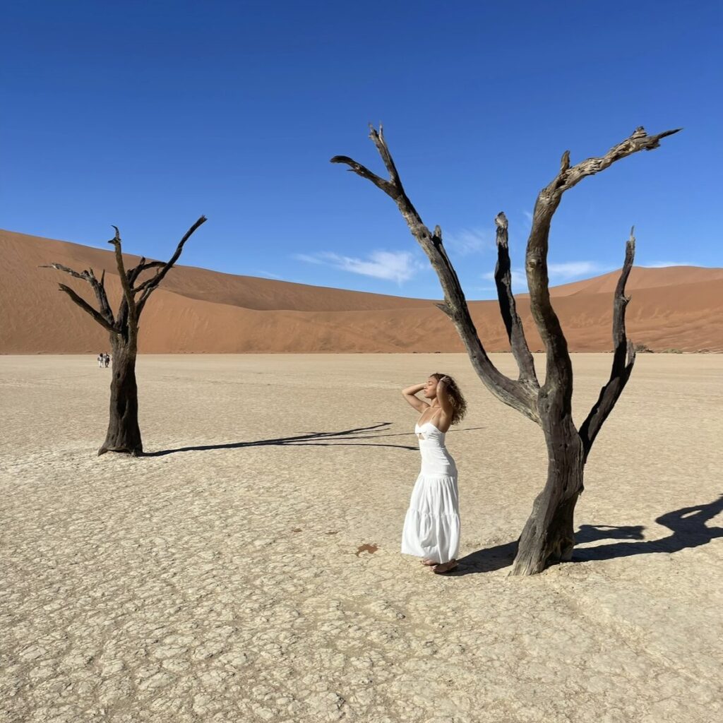 Curly girl in white flowy dress posing next to deadvlei trees in Namibia