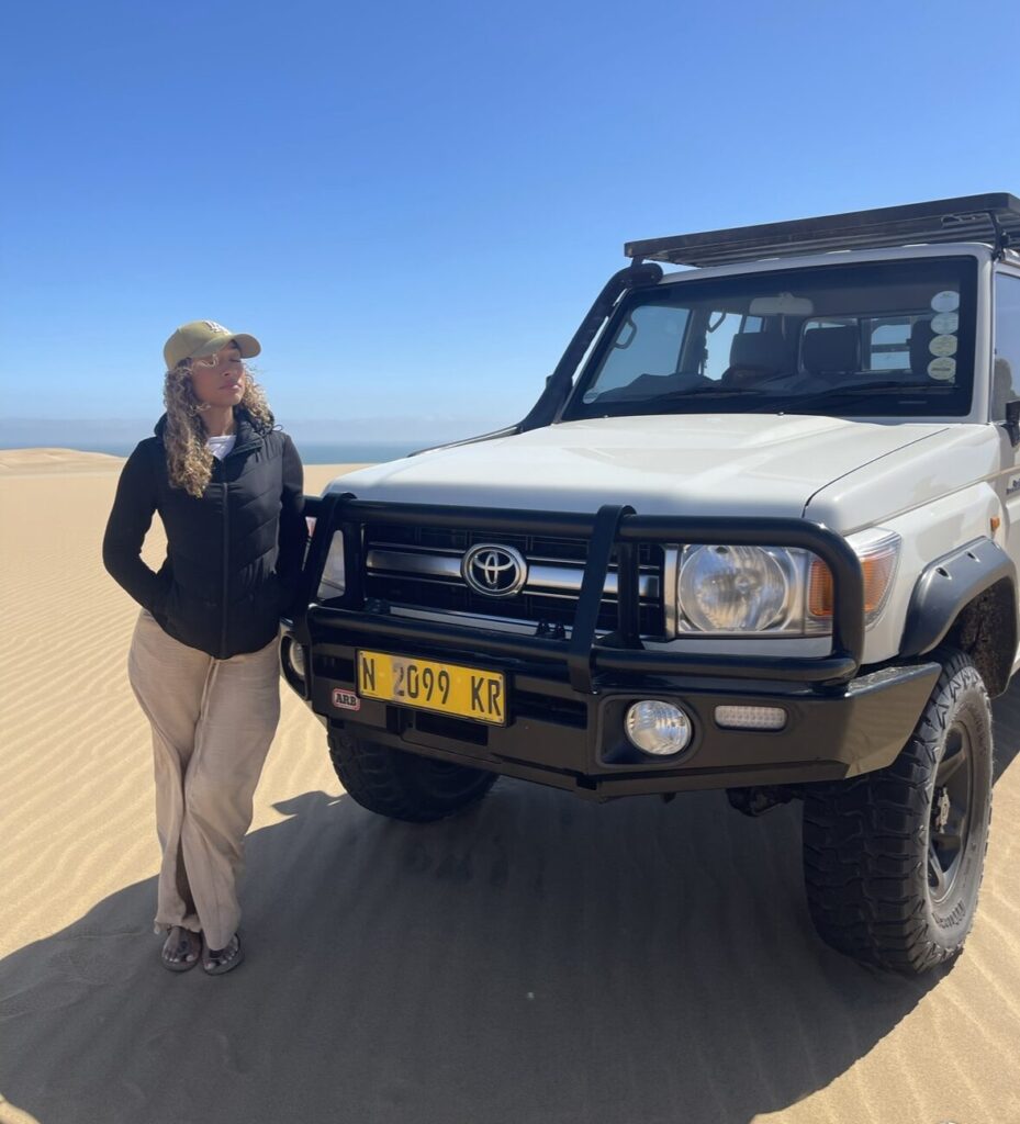 curly haired girl wearing a baseball hat posing next to a 4x4 truck on sand dunes 