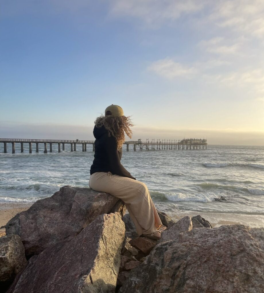 curly haired girl sitting on tall rocks looking ak view of pier in Namibia